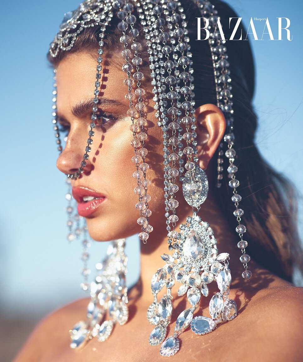 Close-up portrait of a woman adorned with crystal headpiece and earrings against a blurred blue sky for a fashion editorial.