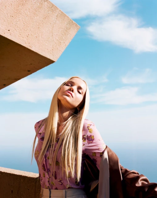 Woman with long blonde hair, eyes closed, wearing a floral shirt, posing outdoors near an architectural concrete structure under a blue sky.