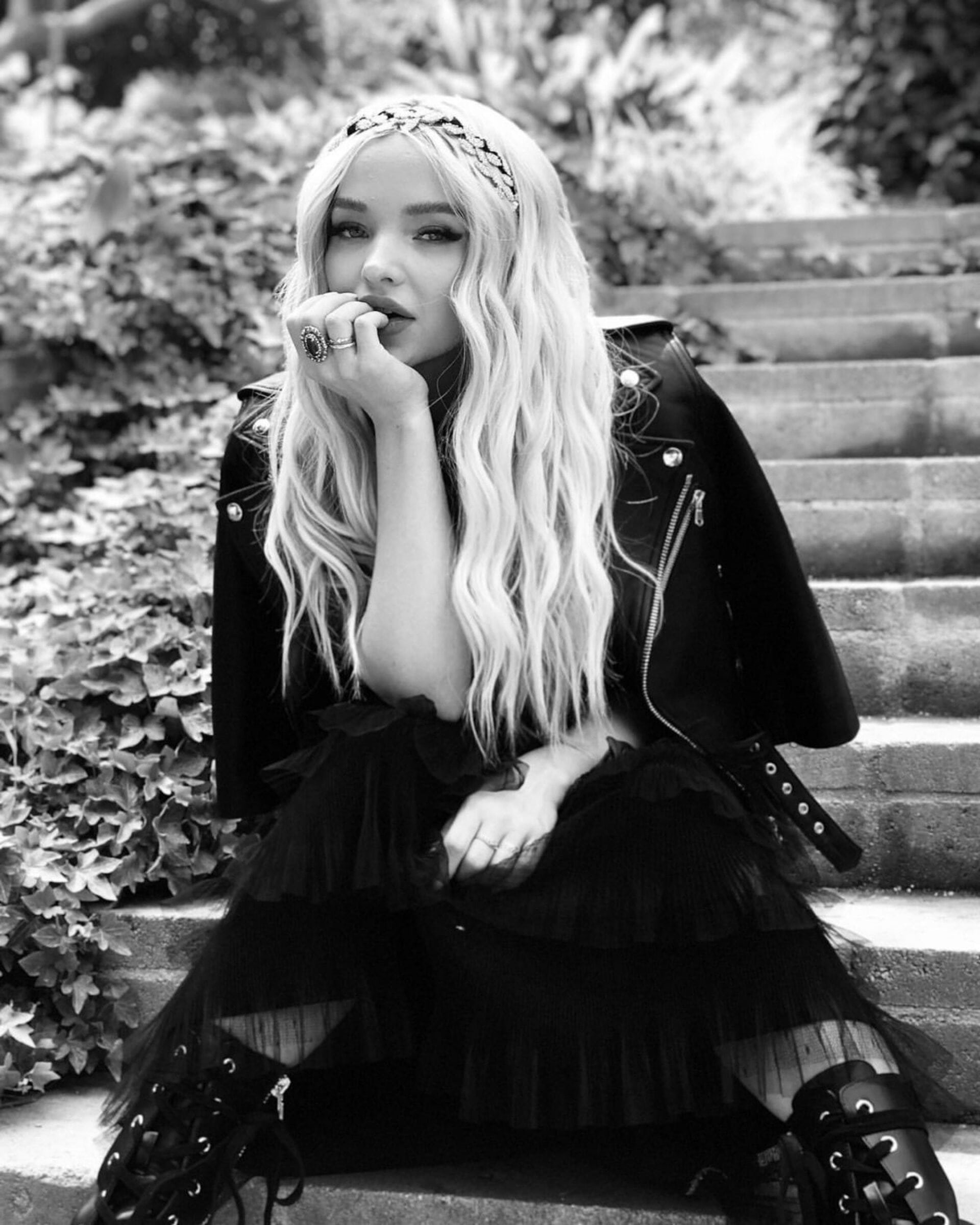 Black and white portrait of an adult female in a layered black dress, headpiece, and leather jacket sitting on stone steps in a garden.