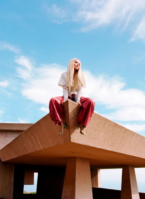 A woman with long blonde hair sits on a building against a bright blue sky with scattered clouds. She is wearing wide red pants.