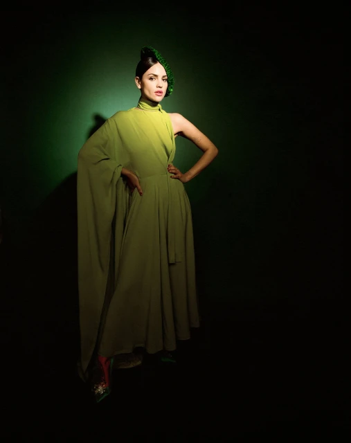 Woman posing in a green dress and unique green headpiece against a dark green backdrop in a studio setting. Full body shot.