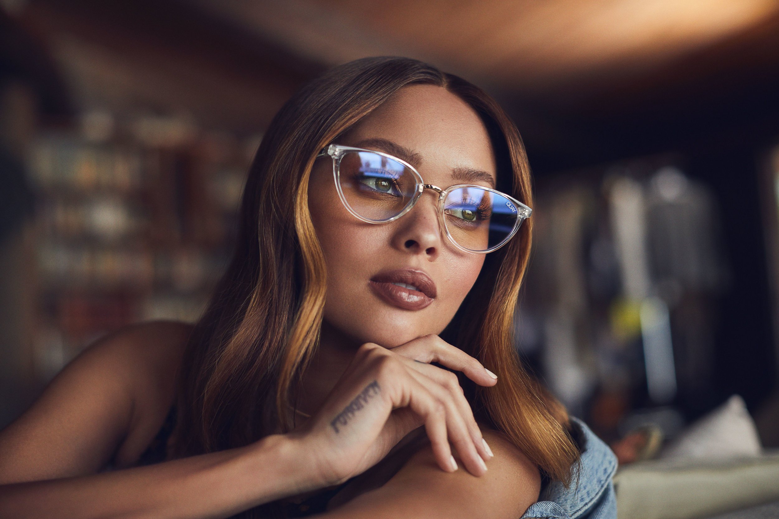 Close-up shot of a stylish adult woman wearing clear-framed glasses, with her hand near her face. Light brown hair frames her face.