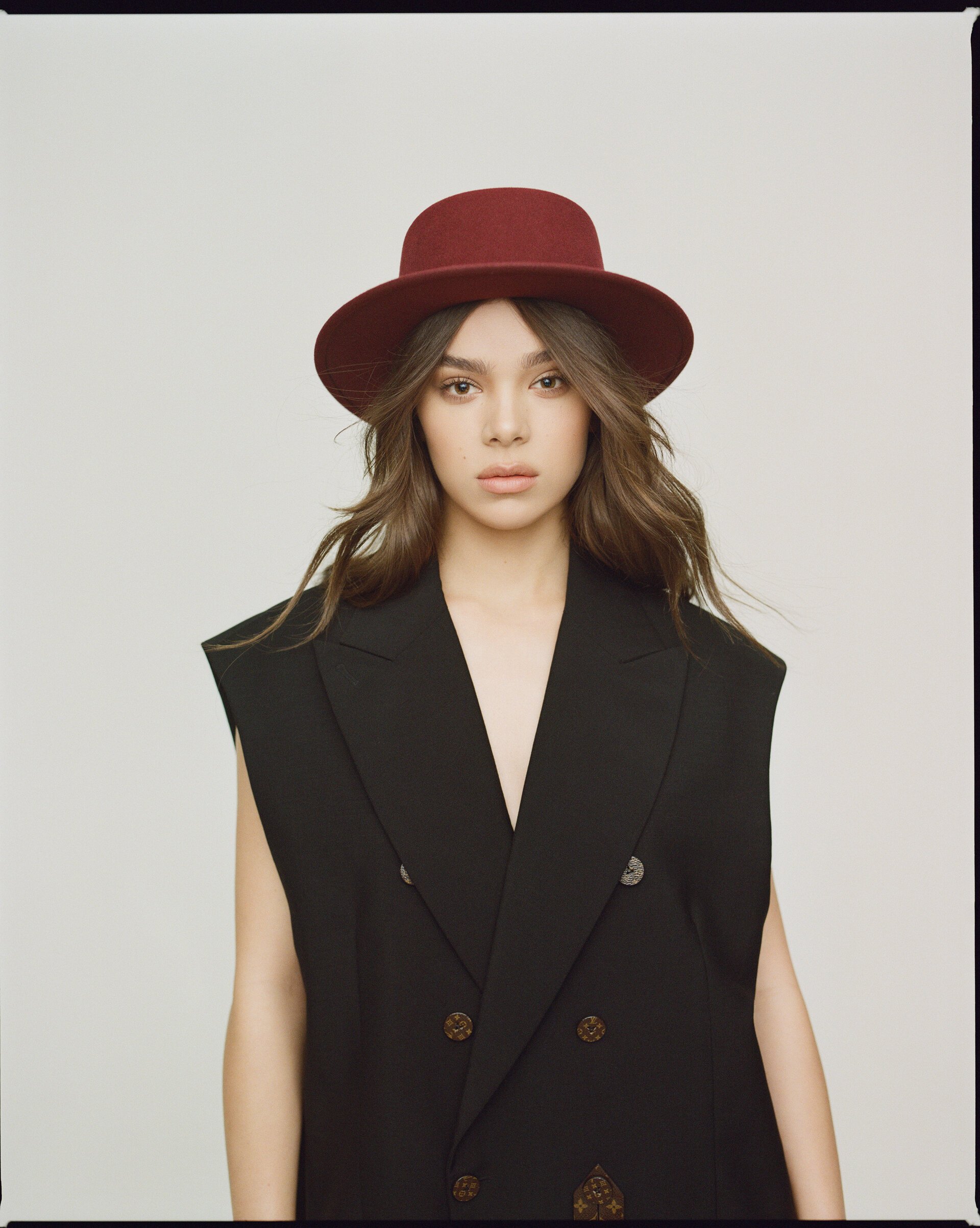 Portrait of a woman with long wavy hair wearing a black double-breasted vest and a maroon hat against a neutral backdrop.