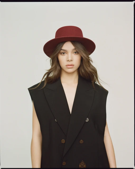 Portrait of a woman with long wavy hair wearing a black double-breasted vest and a maroon hat against a neutral backdrop.