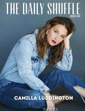 Camilla Luddington wearing a denim shirt and jeans seated against a dark grey seamless backdrop