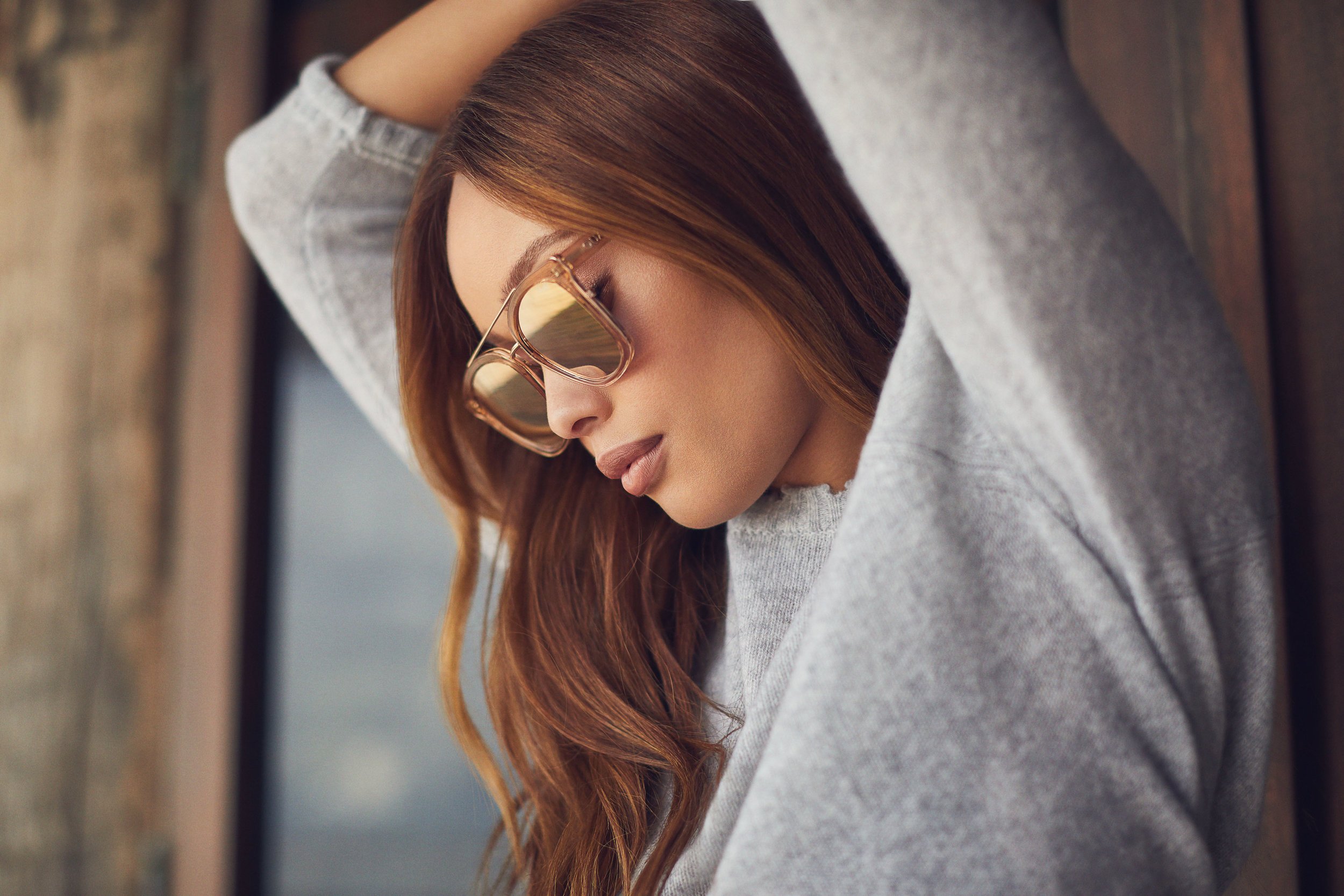 Close-up portrait of a woman wearing tinted sunglasses and a grey knitted sweater with her arms raised