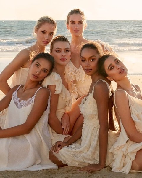 Group of six diverse women posing on a sandy beach in flowing white dresses, soft light, and ocean backdrop. Fashion, beauty, lifestyle.