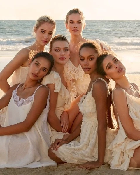 Group of six diverse women posing on a sandy beach in flowing white dresses, soft light, and ocean backdrop. Fashion, beauty, lifestyle.