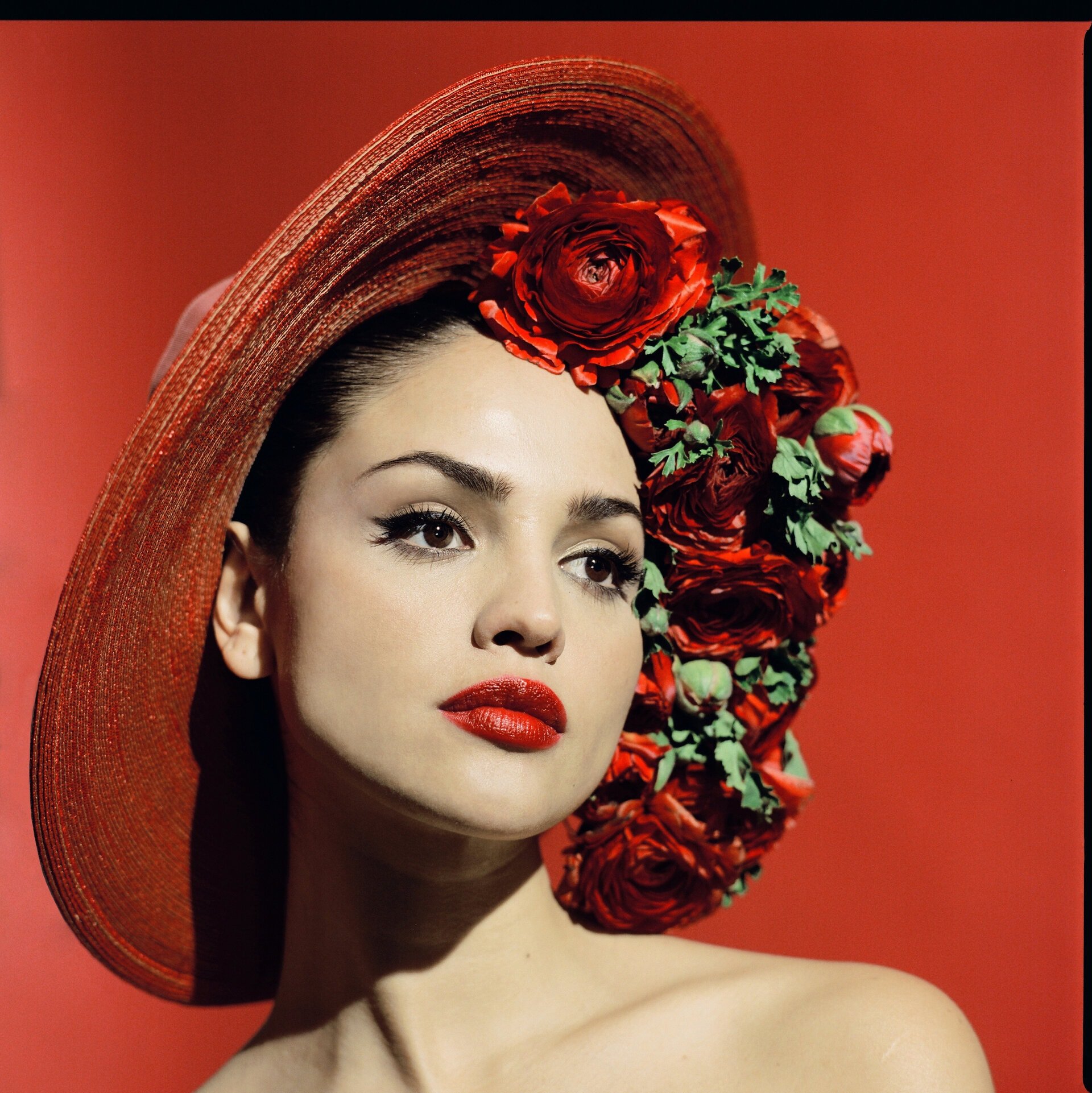 Woman with a red hat decorated with roses, posing against a matching red backdrop in a fashion or beauty editorial-style portrait.