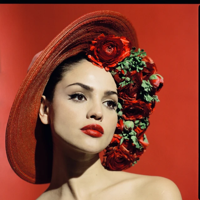 Woman with a red hat decorated with roses, posing against a matching red backdrop in a fashion or beauty editorial-style portrait.
