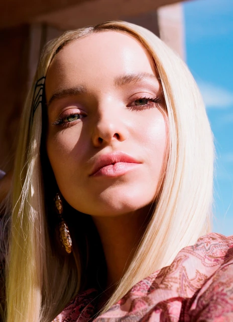 Close up portrait of a blonde woman with olive skin wearing gold earrings and a pink patterned top in an outdoor setting with blue sky.