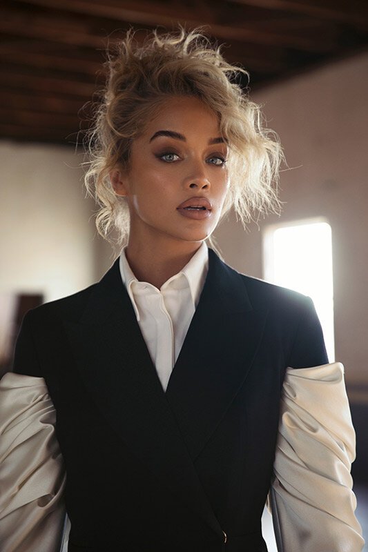 Close-up portrait of a fair-skinned woman with blonde curly hair in a black blazer and white shirt, posing in a sunlit interior space.