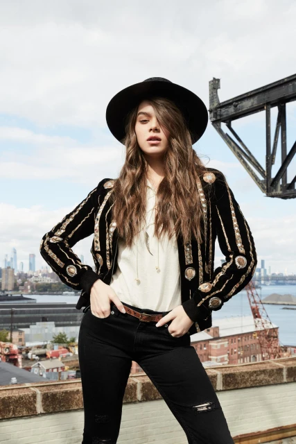 A young woman with long hair stands on a rooftop wearing a black hat, jacket, jeans, and boots, with a city skyline in the background.