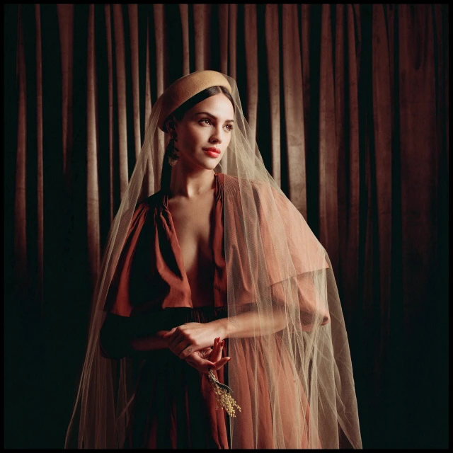 Indoor portrait of a young adult woman with a beige hat and veil, wearing a copper dress, standing in front of a draped curtain backdrop.