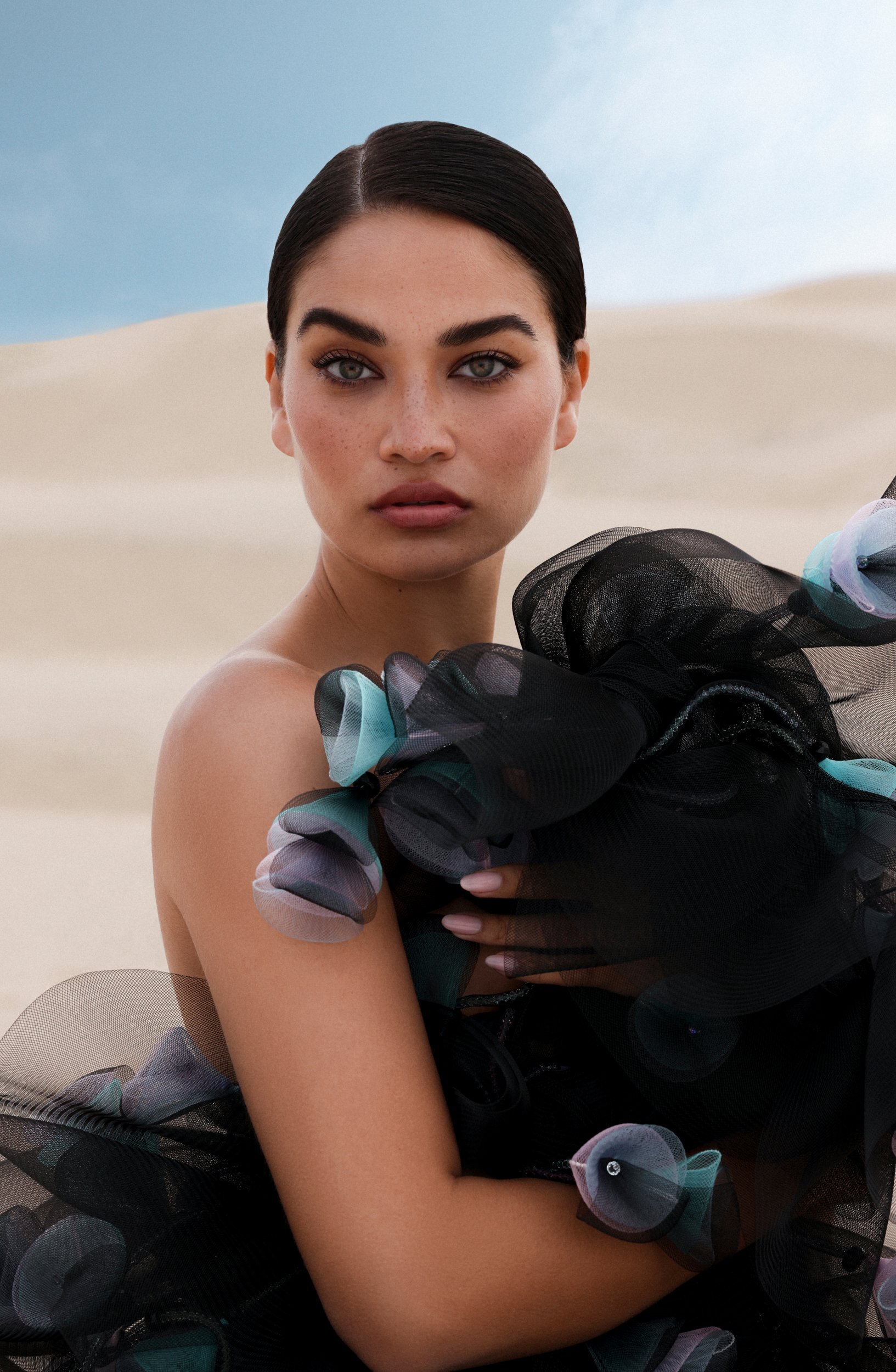 Portrait of a woman in a ruffled tulle dress posing against a sand dune under a blue sky, creating a striking contrast of fashion and nature.