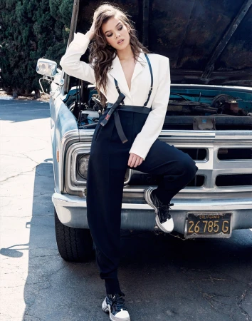 Hailee Steinfeld poses in white blazer and black pants leaning on an old pickup truck. Automotive fashion editorial photoshoot.