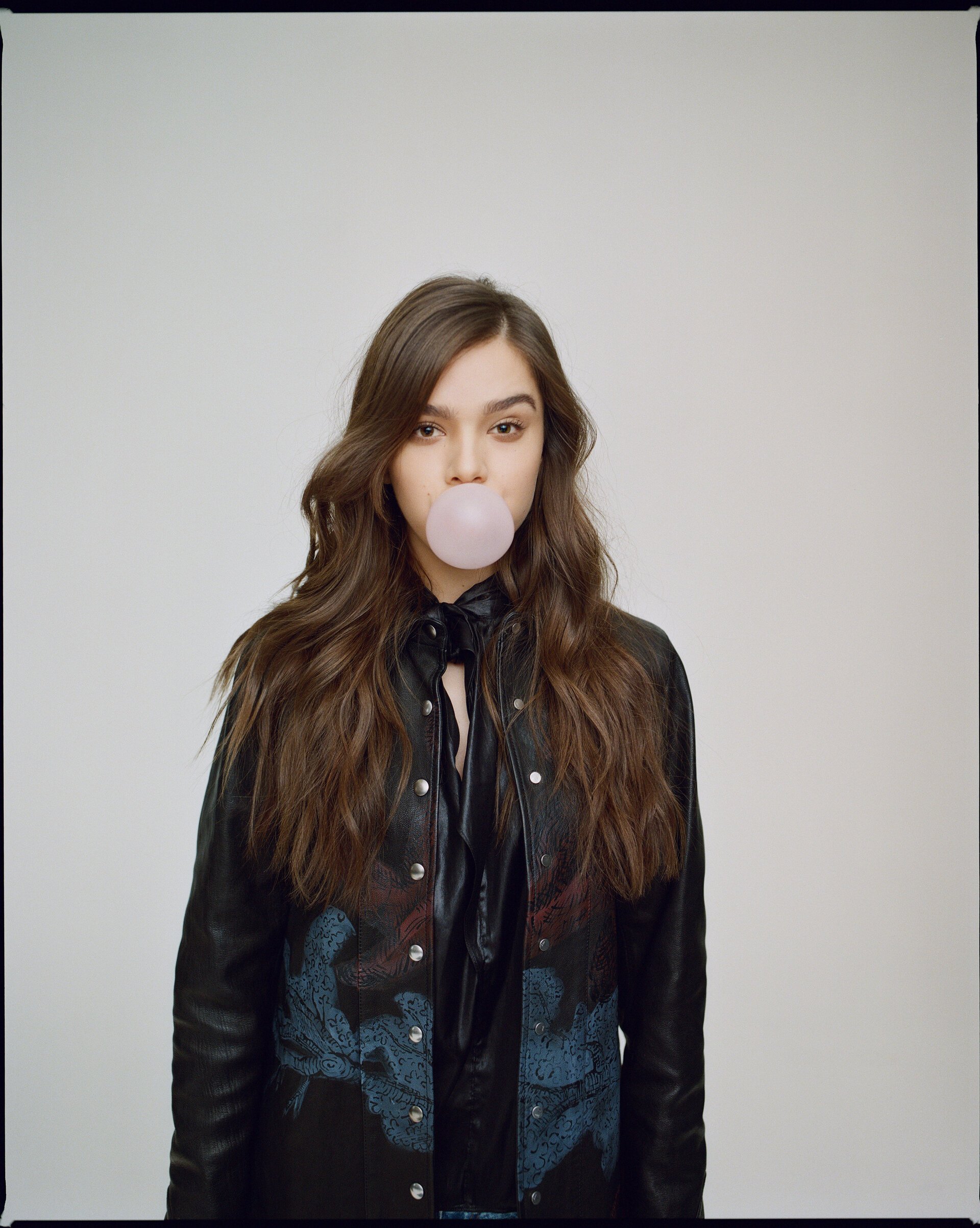 A young woman with long brown hair blows a pink bubble gum bubble in a studio shot. She wears a black leather jacket.