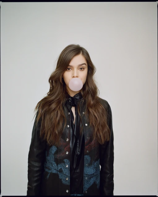 A young woman with long brown hair blows a pink bubble gum bubble in a studio shot. She wears a black leather jacket.