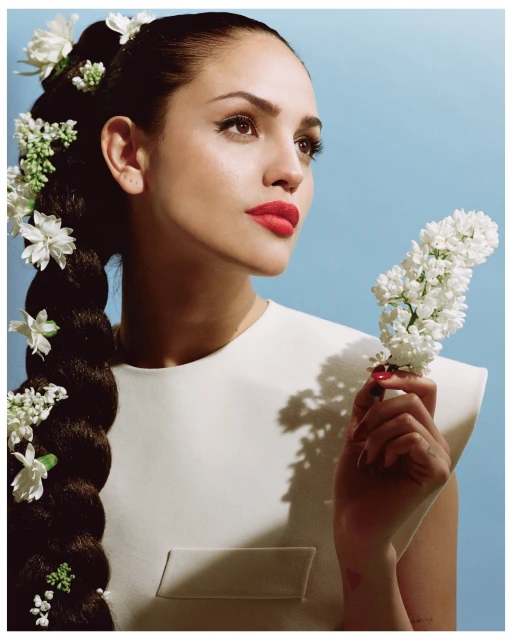 Portrait of a woman with a long braided hairstyle adorned with white flowers, wearing a white dress, and holding a flowering branch.