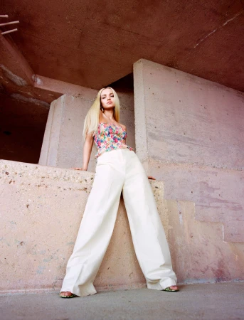 Blonde woman in a floral top and white pants leaning against a concrete wall in an outdoor urban setting with soft lighting.