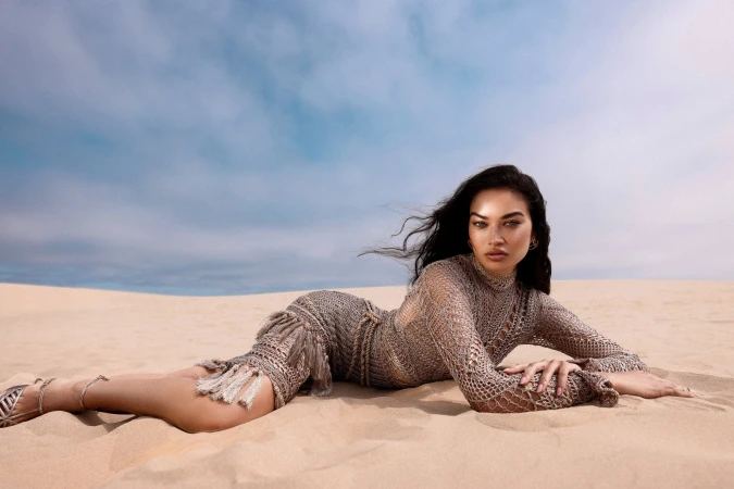 Model with long dark hair lying on sand dunes wearing a beige crocheted fringe dress against an open sky.