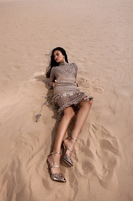 Fashion shot of a woman in a beige dress and high heels lying on sand in a desert landscape. Warm tones and shallow depth of field.