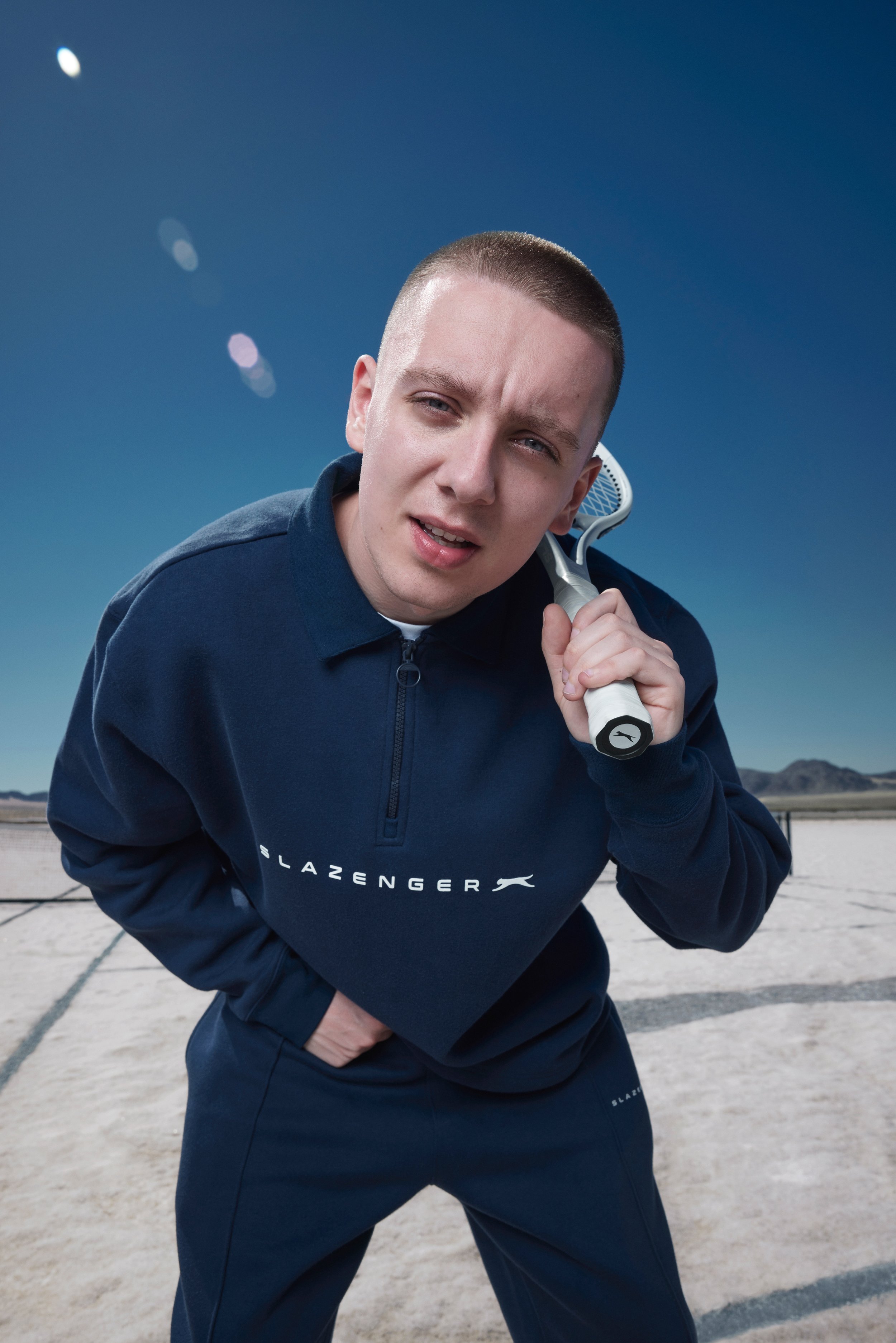 Adult male wearing a navy blue Slazenger tracksuit in an outdoor setting, holding a tennis racket up to his neck.