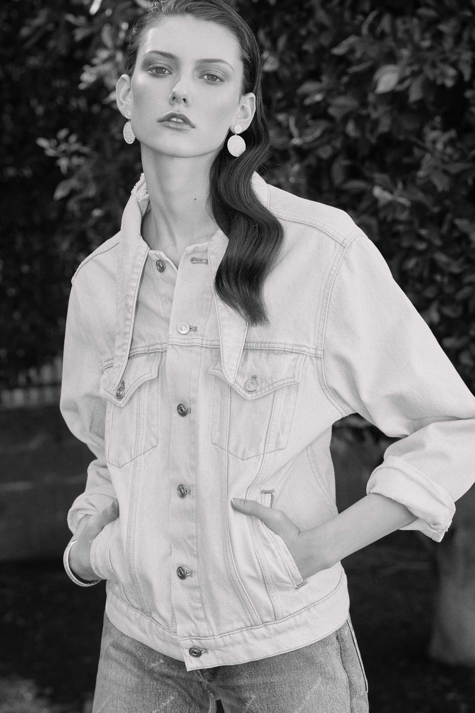 Monochrome fashion portrait of a young woman in a denim jacket with a soft-focus backdrop of foliage, highlighting style and texture.