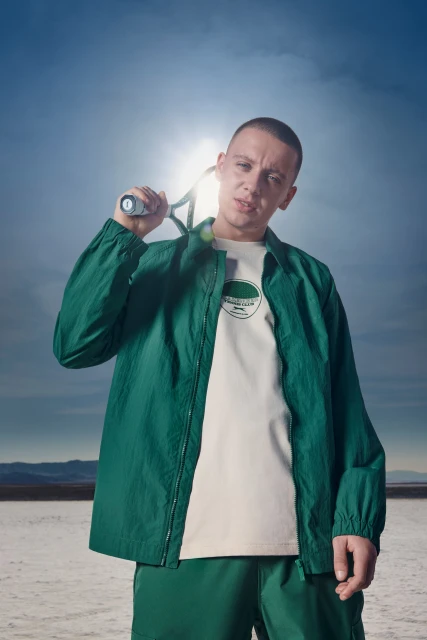 Adult male poses outdoors in a green jacket and shorts with a tennis racket over his shoulder, standing on a salt flat.