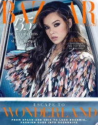 Hailee Steinfeld seated inside a car wearing a feathery textured top with dark eye makeup and styled dark wavy hair.