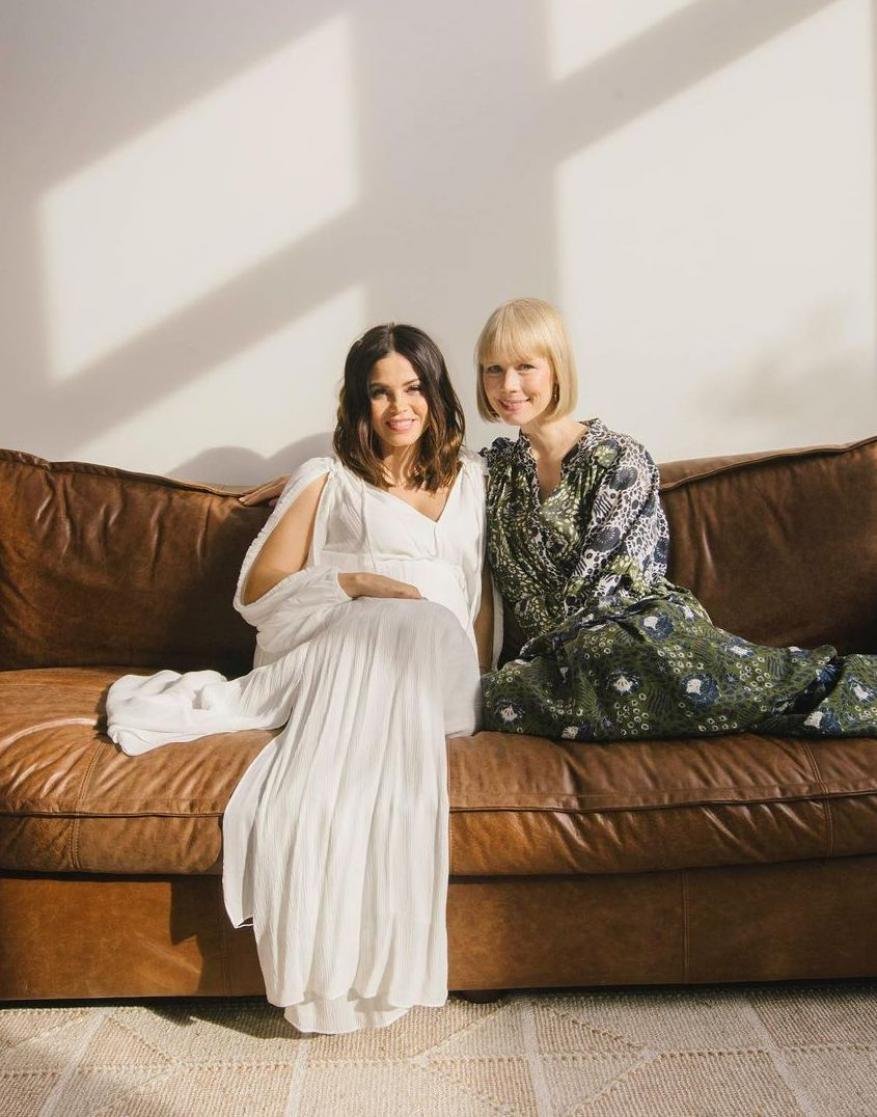 Two women pose on a brown leather couch in a brightly lit room. One is wearing a white dress and the other a patterned dark green dress.