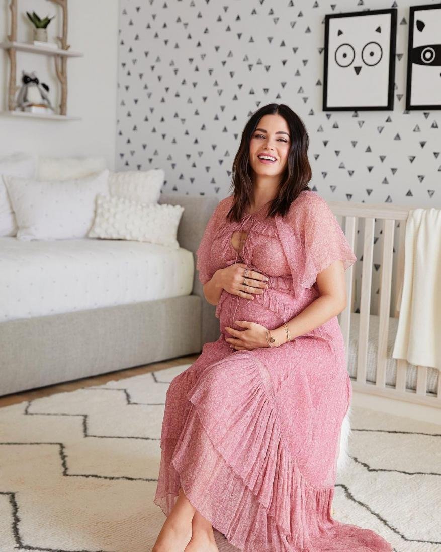 Pregnant woman in a pink dress sits in a nursery with a crib and triangular patterned wallpaper, looking at the camera and smiling.