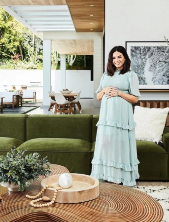 A pregnant woman in a light blue dress stands in a modern living room with a green velvet sofa and a patterned wooden coffee table.