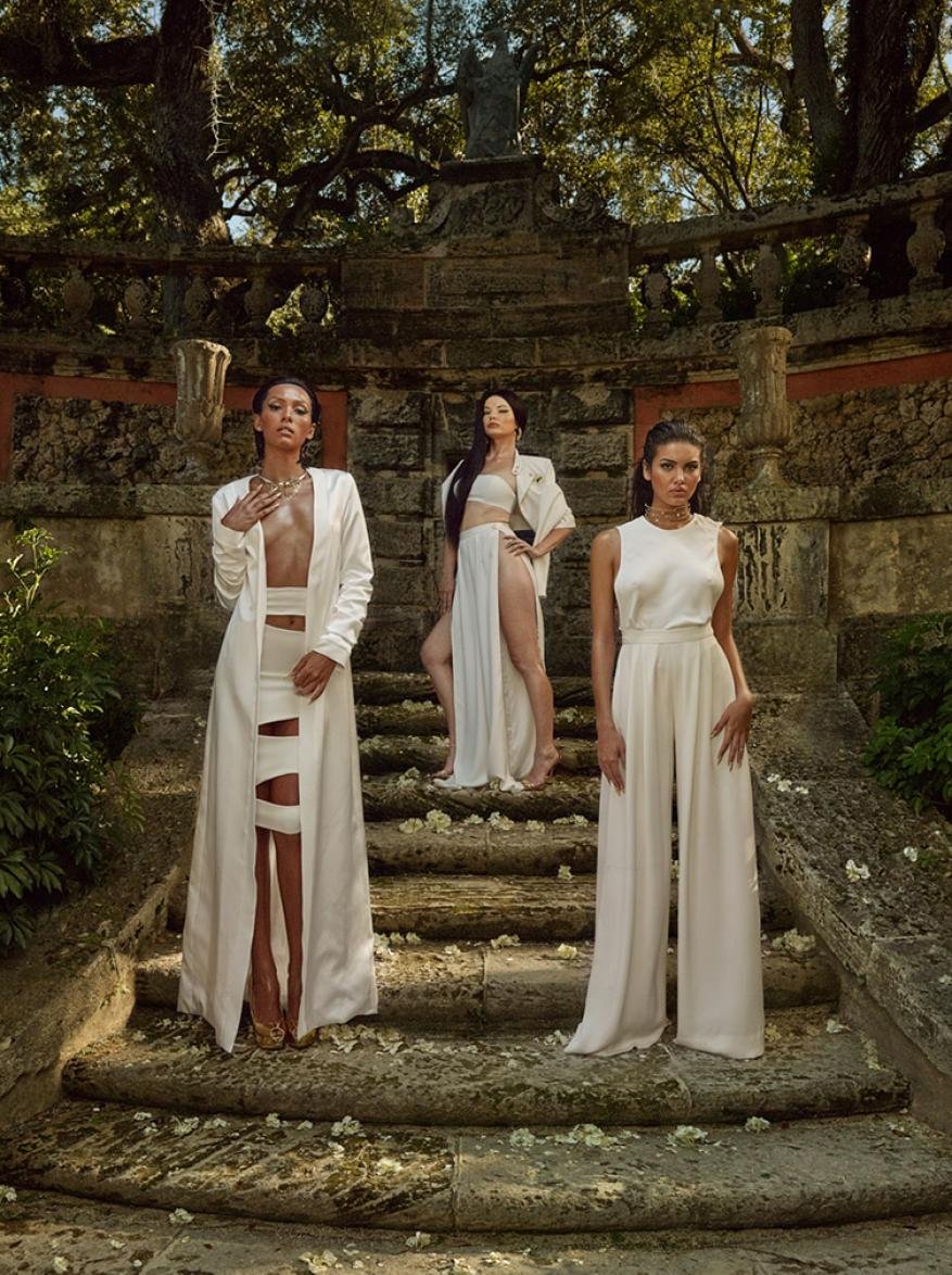 Three fashion models in white outfits pose on stone steps surrounded by greenery in an outdoor setting, showcasing elegant attire.
