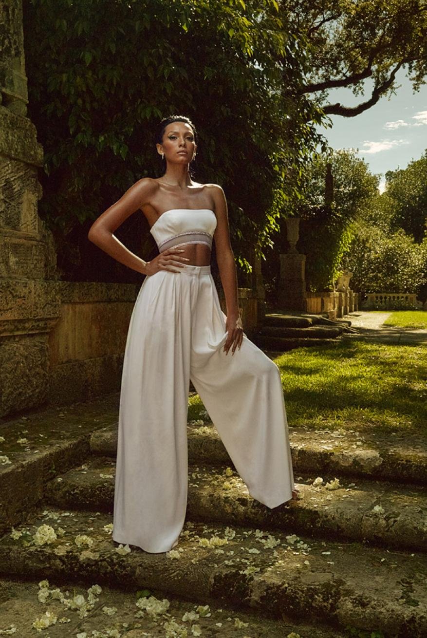 Elegant woman in a matching white crop top and palazzo pants poses on steps with scattered petals in a lush garden setting.