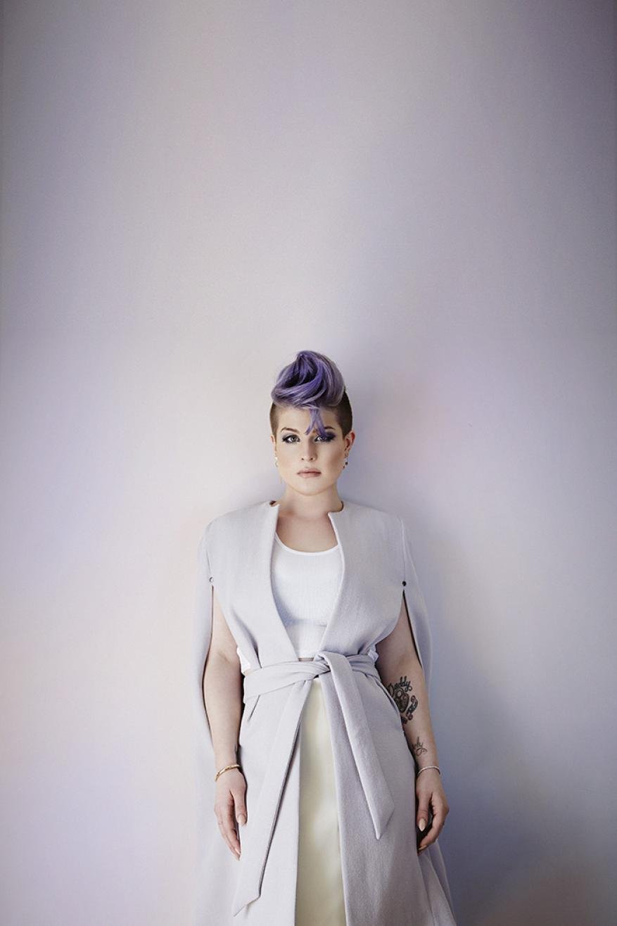 Portrait of adult woman with purple hair, wearing a light-colored dress with tie belt, standing indoors against a neutral wall.