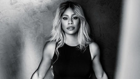 Portrait of Laverne Cox with blonde-highlighted hair in a dark sleeveless top against a textured wall