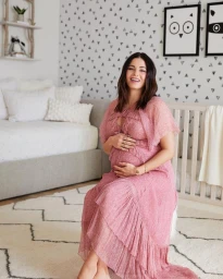Pregnant woman with dark hair wearing a pink ruffled dress sitting in a nursery with patterned wallpaper