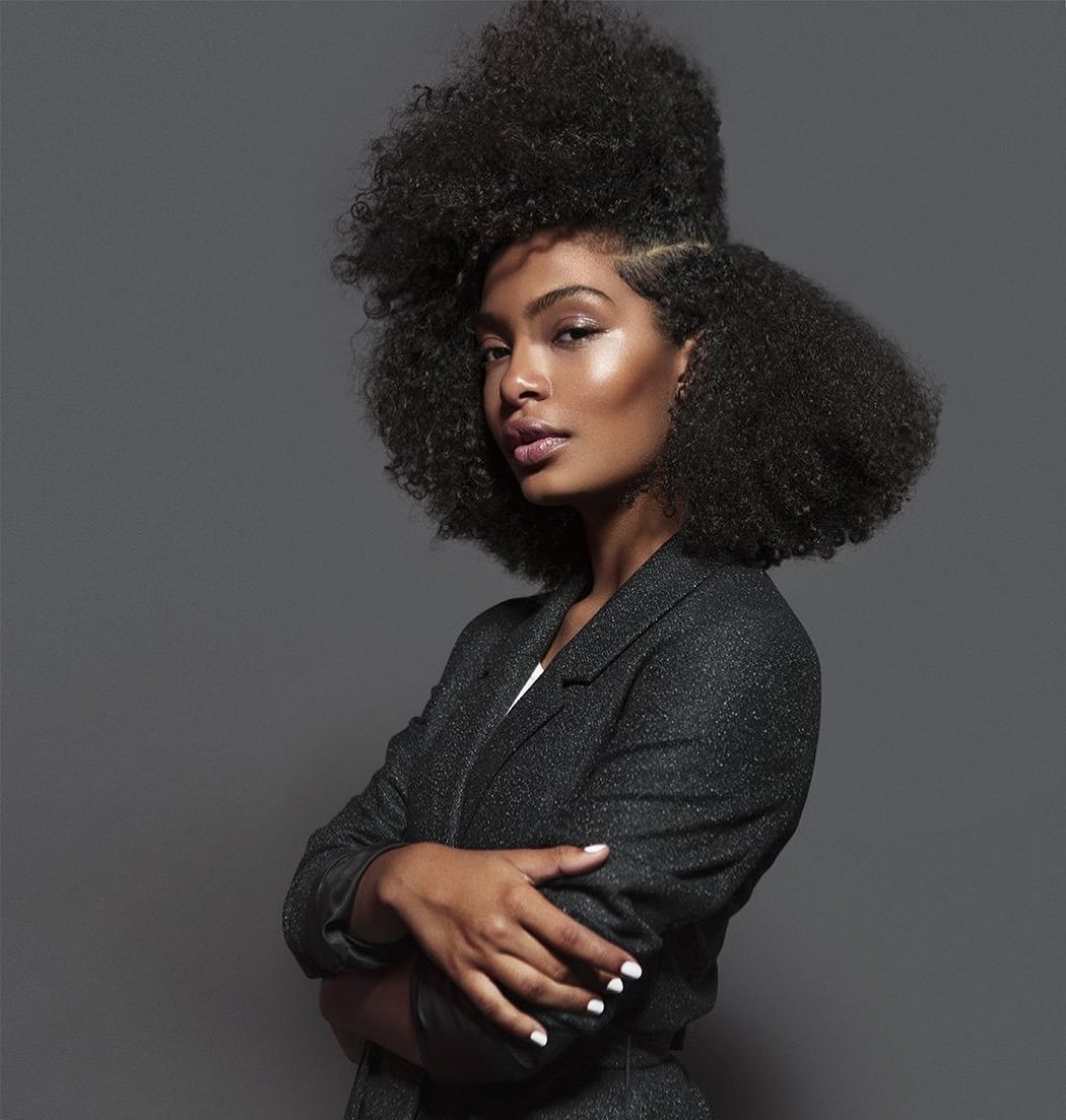 Portrait of a Black woman with an afro, wearing a blazer with folded arms, against a gray background. Beauty and fashion concept.