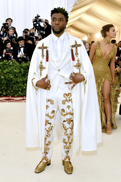 Chadwick Boseman in a white suit and cape with gold embellishments, attending an event with photographers and another person in the background.