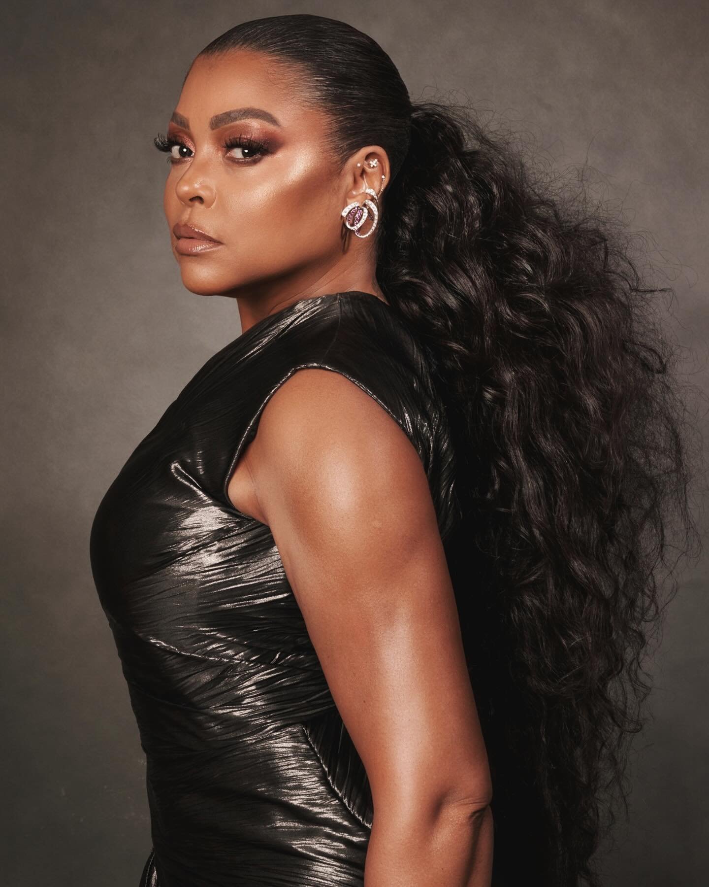 A portrait of an adult woman with dark skin and dark wavy hair in a high ponytail, wearing a shiny black dress against a grey background.