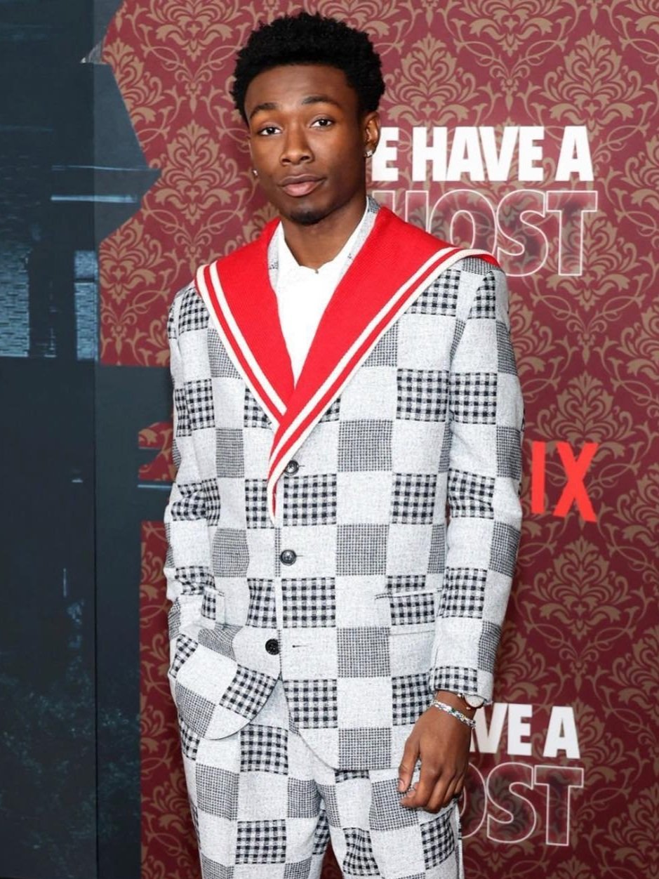 A young African American man in a patterned suit stands in front of a backdrop with a promotional logo for a film or entertainment event.