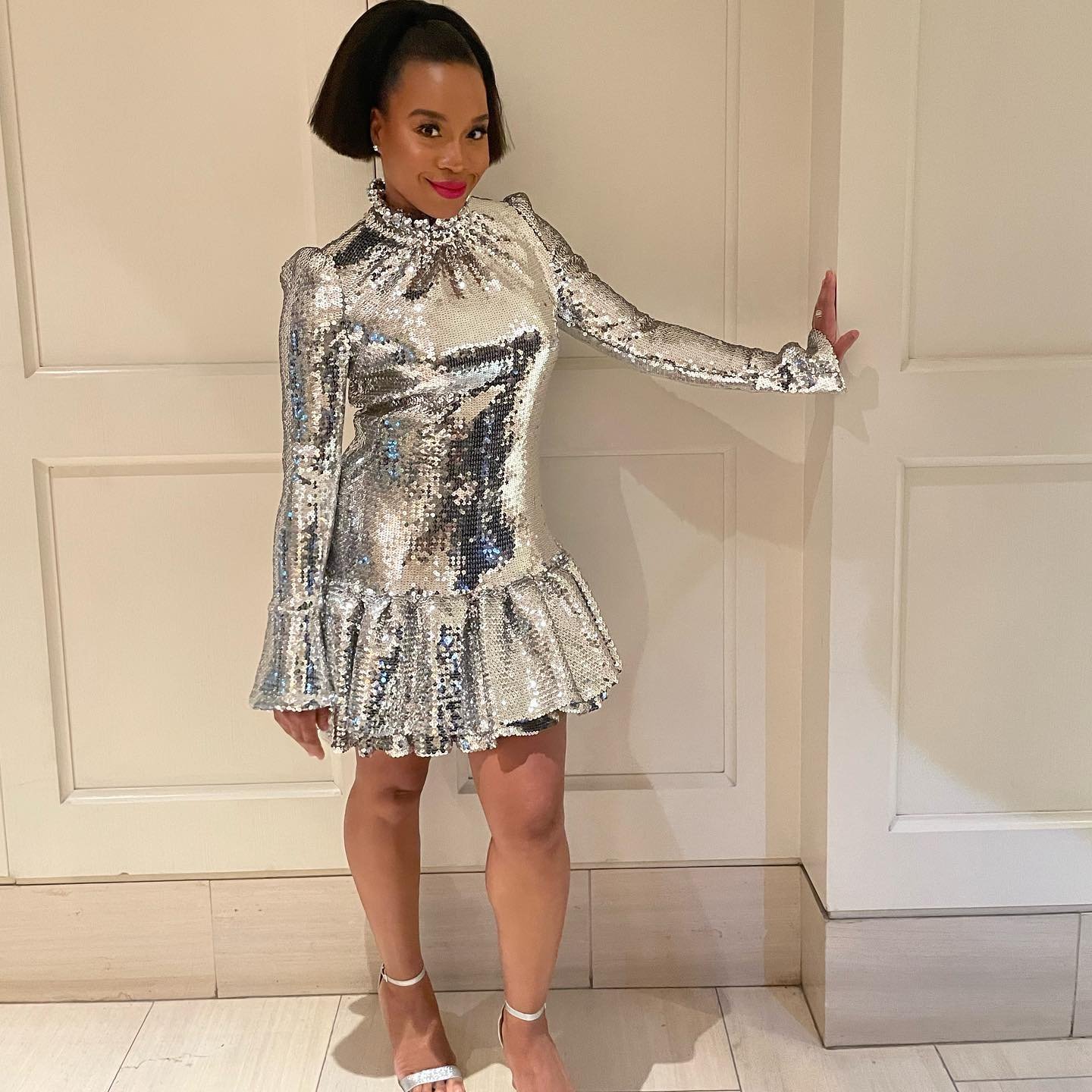 Woman in sequin mini dress with high collar and ruffle hem poses in front of paneled wall with a hand resting on the trim.
