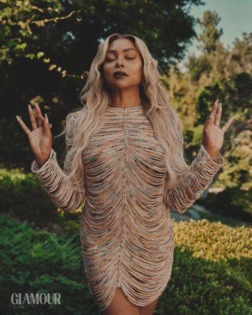 Glamour shot of an adult woman with long blonde hair, wearing a tan fringe dress, with her eyes closed and raised hands in an outdoor setting.