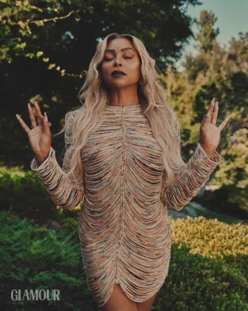 Glamour shot of an adult woman with long blonde hair, wearing a tan fringe dress, with her eyes closed and raised hands in an outdoor setting.