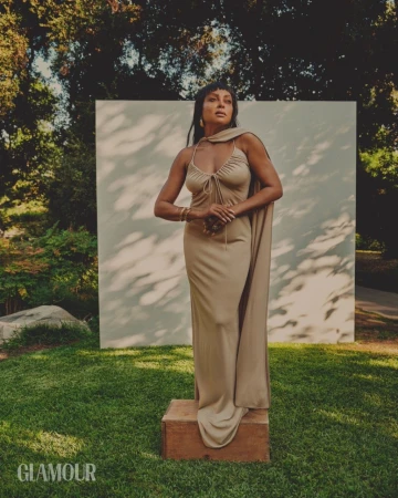 Woman in a draped beige halterneck dress standing on a wooden box outdoors with green foliage and a white backdrop panel.