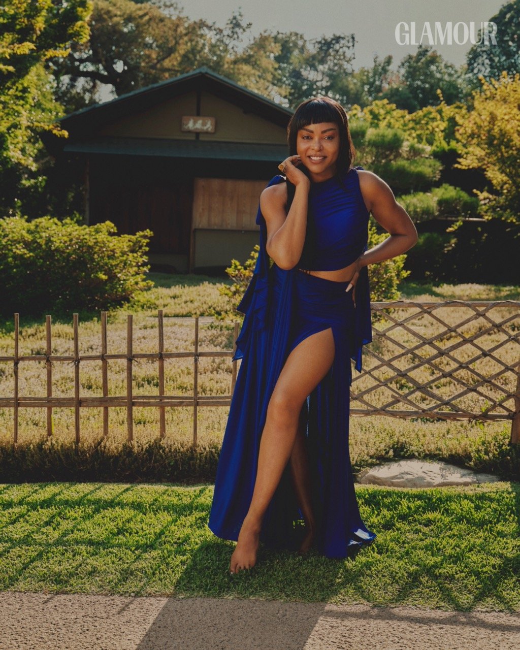 Glamorous woman in a blue high-slit dress poses barefoot in a lush garden setting with a traditional Japanese-style house.