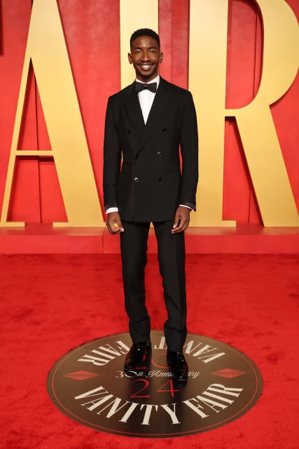Full-length portrait of a Black man in a black tuxedo and bow tie, standing on a red carpet with the words 'Vanity Fair' in the background.