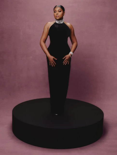 Elegant adult woman in a floor-length black halter dress and diamond jewelry stands on a circular platform against a neutral backdrop.