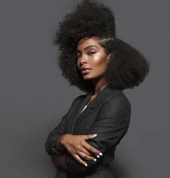 Portrait of Yara Shahidi with voluminous curly hair wearing a dark blazer against a grey backdrop